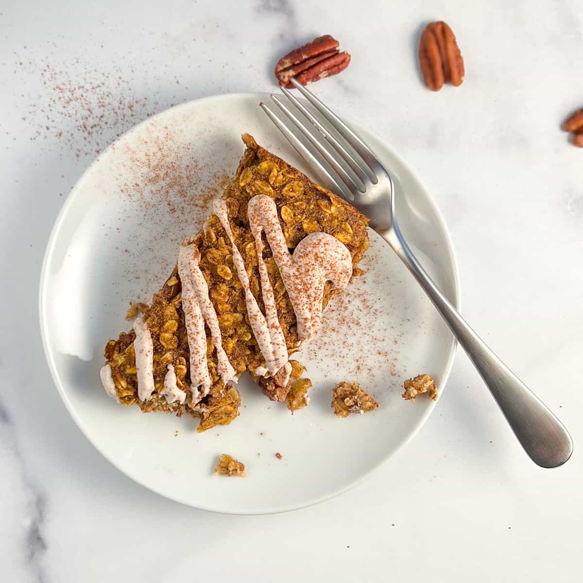 top view close up of a pumpkin oatmeal bar with sweet cashew cream drizzled on top on a round white plate with fork on the side and loose pecans scattered on a marble surface