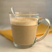side view close up pumpkin spice milk latte in a clear glass mug with a stainless teaspoon sticking out. yellow napkin blurred in the background