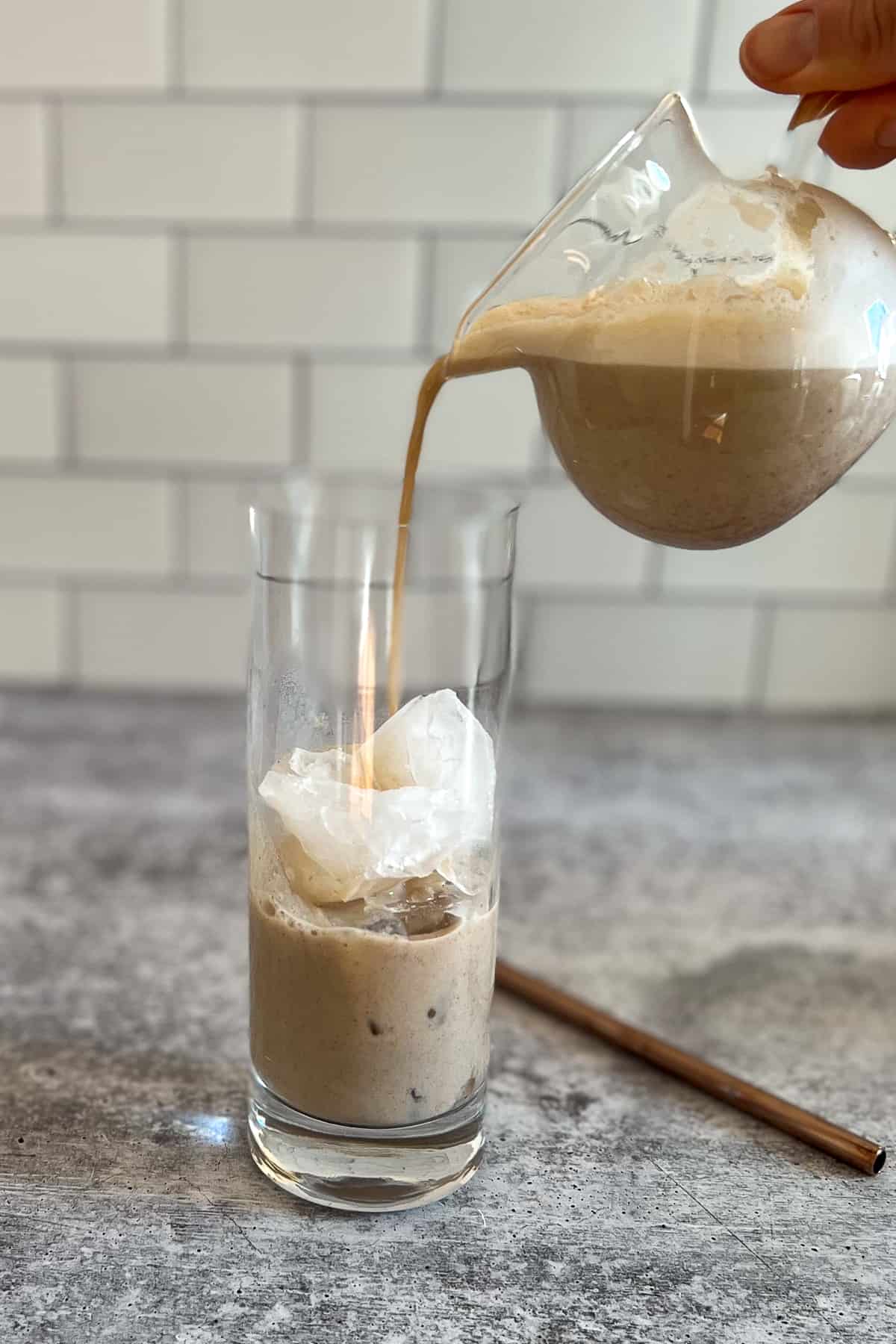side view of a woman's hand holding a small pitcher of iced pumpkin spice latte being poured into a tall glass with ice. metal straw on the side