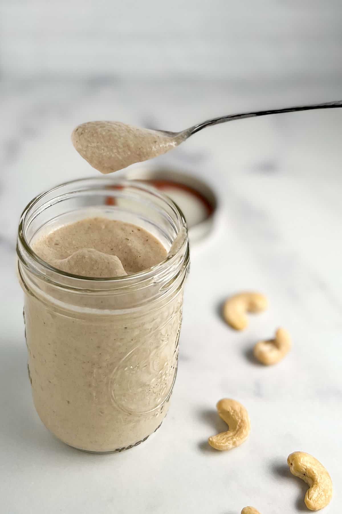 top side view of sweet cashew cream sauce in a glass mason jar with spoon being dipped. Loose cashews blurred in the background.