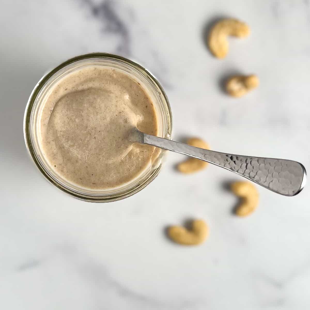 top view close up of sweet cashew cream in a glass mason jar with a spoon sticking out. loose cashews blurred in the background