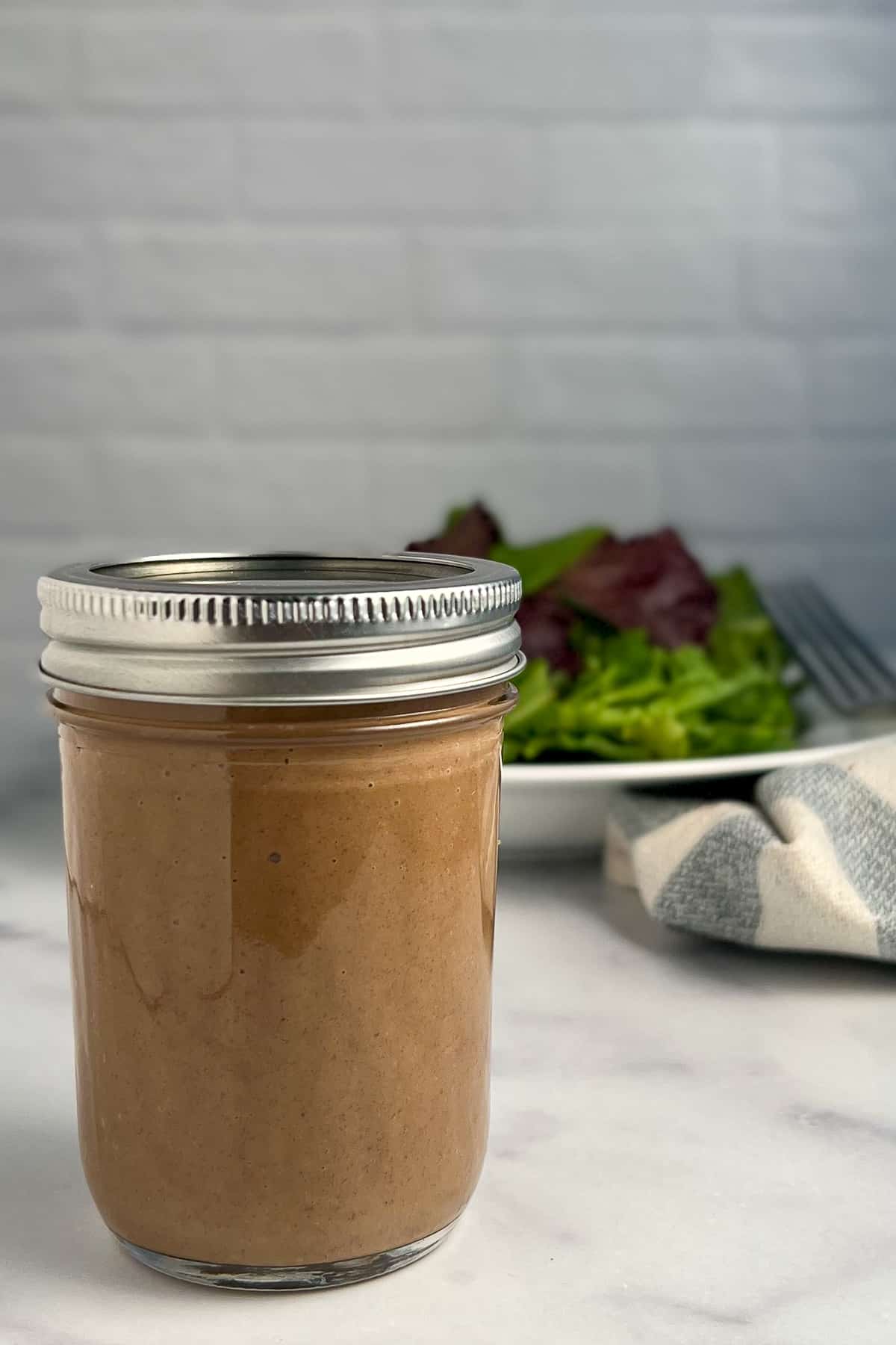 side view close up of a mason jar with balsamic vinaigrette dressing and mixed green salad in a white bowl blurred in the background