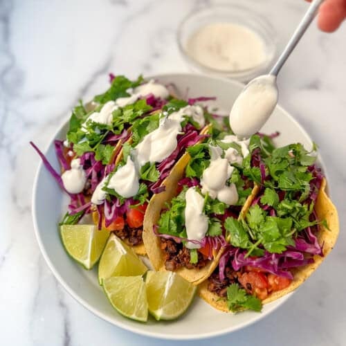 Four black bean tacos; spoon drizzling cashew cream sauce on top.