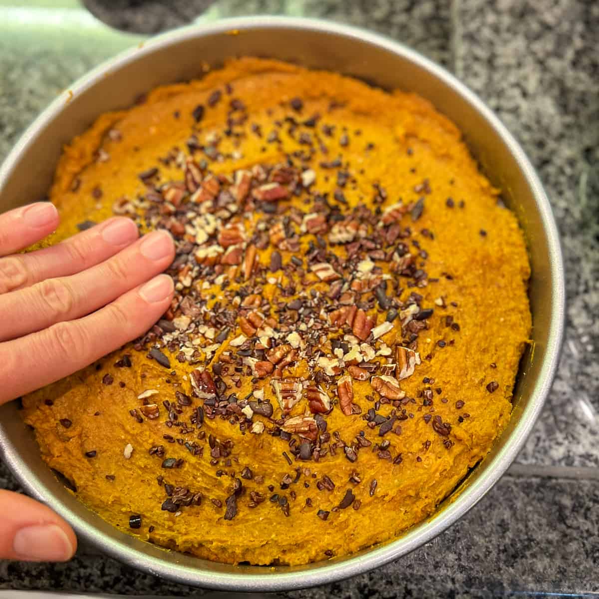 A woman's hand pressing the chopped nuts and cacao nibs into the unbaked crustless vegan sweet potato pie.