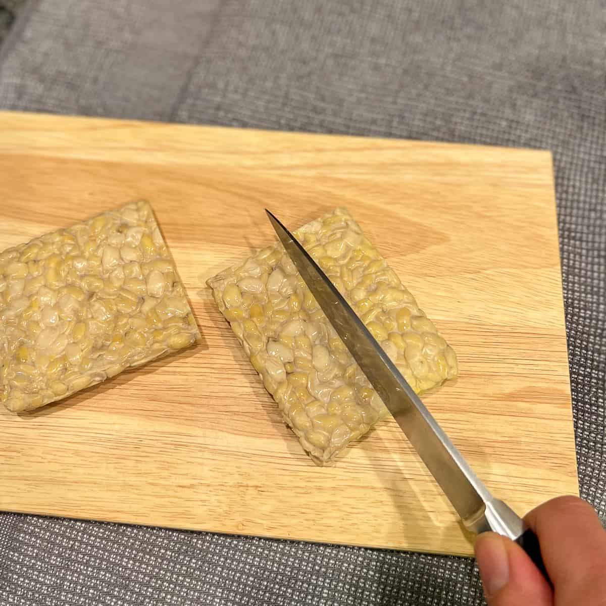 a block of tempeh on a cutting board with a woman's hand holding the knife cutting it into quarters