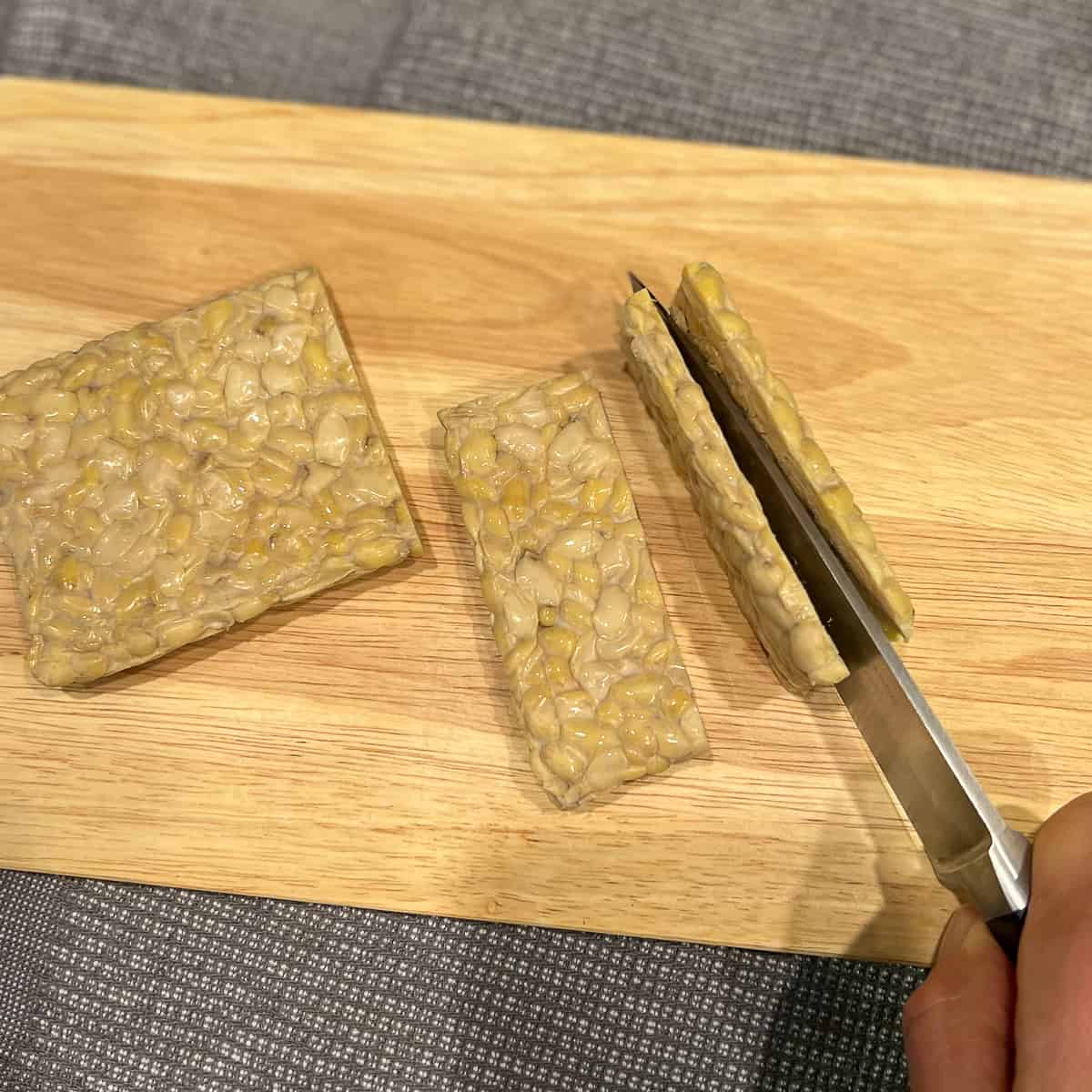 a block of tempeh on a cutting board with a woman's hand holding the knife cutting it in half down the narrow side