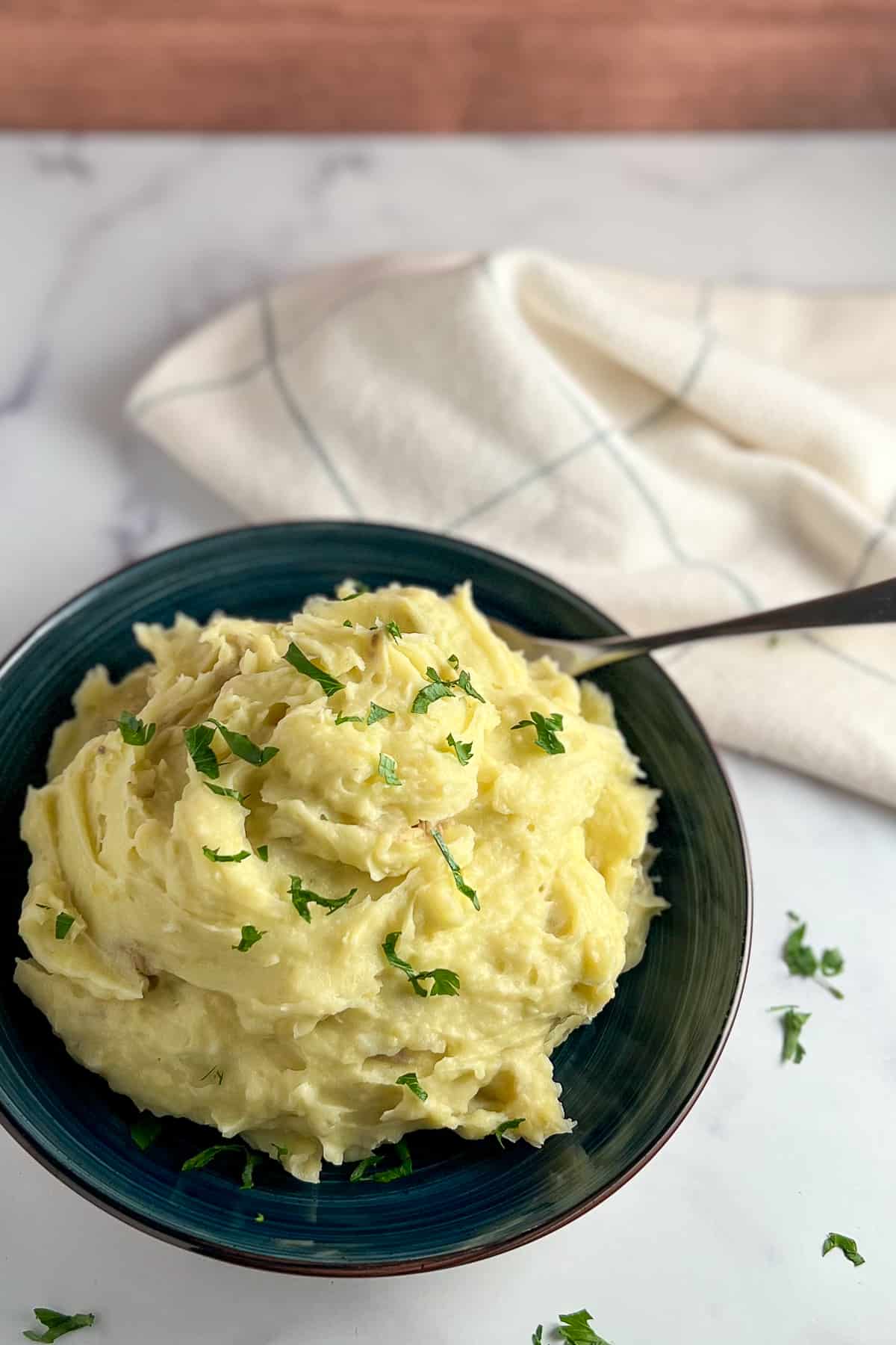 top side view of a bowl of mashed potatoes with fresh chopped parsley and kitchen towel blurred in the background.