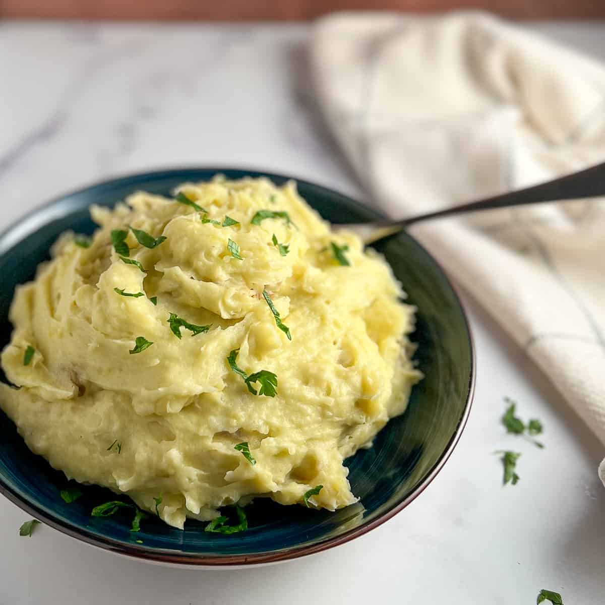 side view of mashed potatoes with fresh chopped parsley in a dark blue bowl with napkin in background