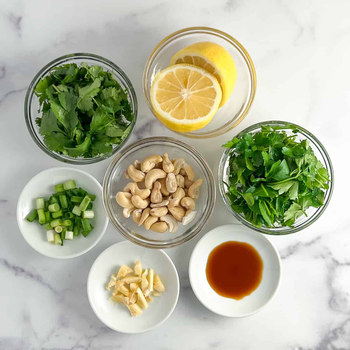top view of key ingredients used in Easy Green Goddess Dressing on a marble surface. cilantro, parsley, lemon juice, cashews, green onion, garlic, coconut aminos.