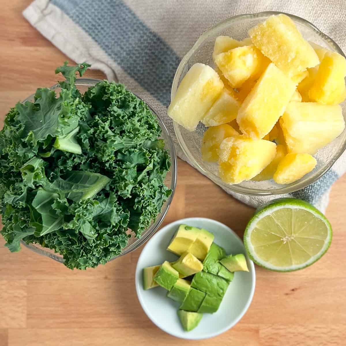 top view of ingredients for kale smoothie: kale, frozen pineapple chunks, avocado, lime