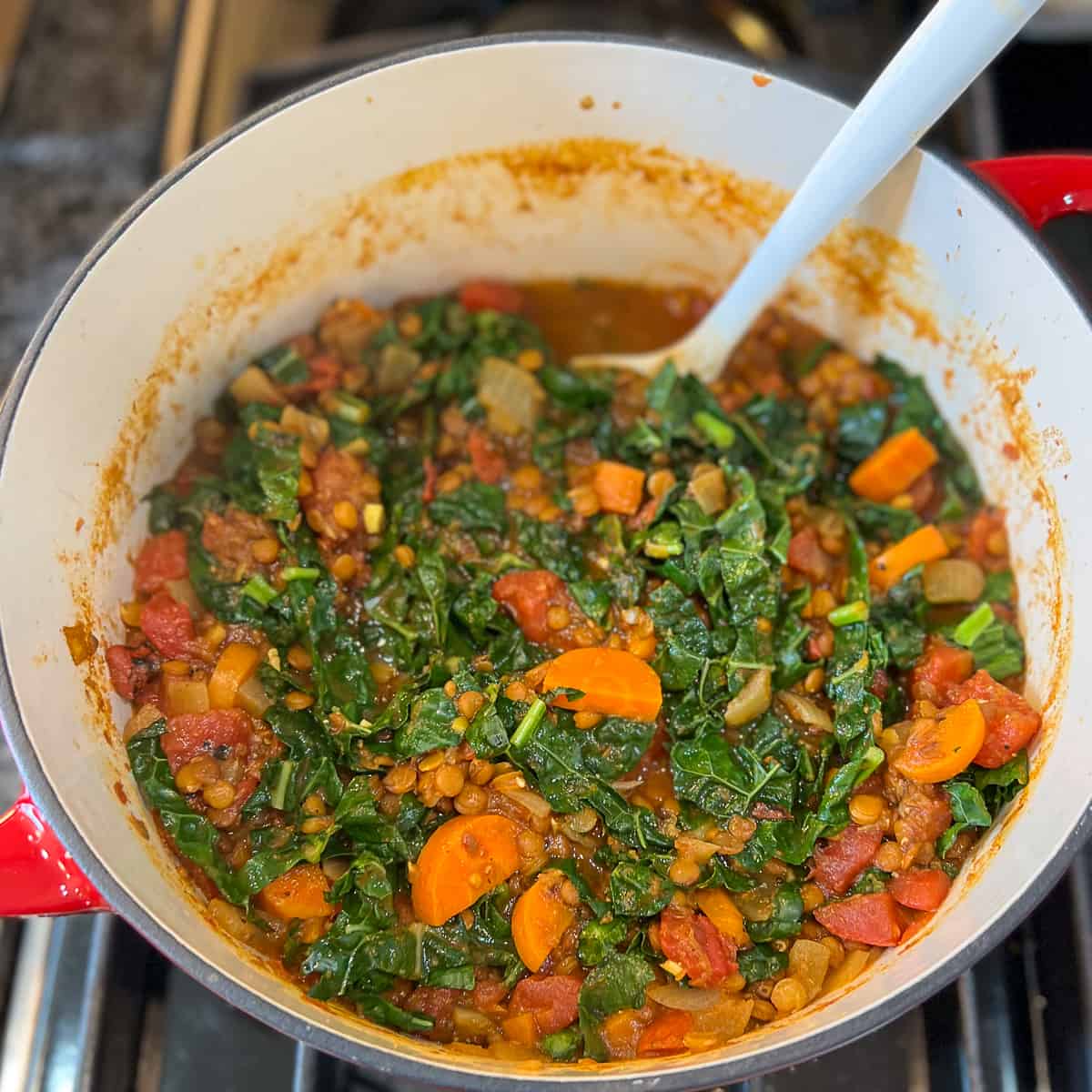 top view close up of a pot of lentil soup with kale, carrots, tomatoes and cooking spoon inside the pot