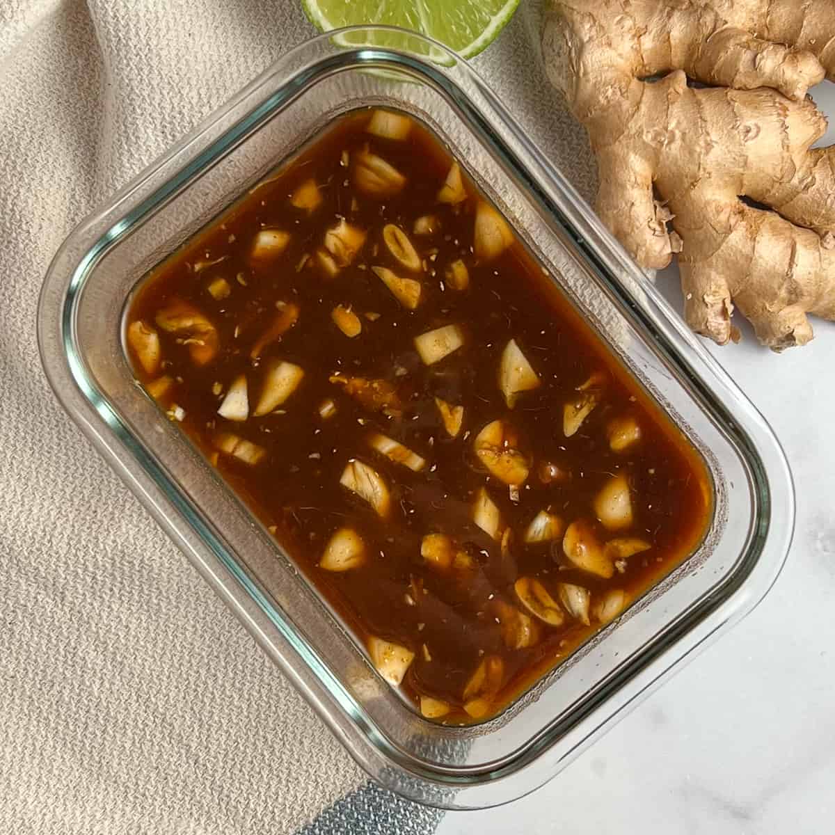 top view close up of stir fry sauce with chunks of garlic in a rectangular glass container; fresh ginger root and lime wedge on the side
