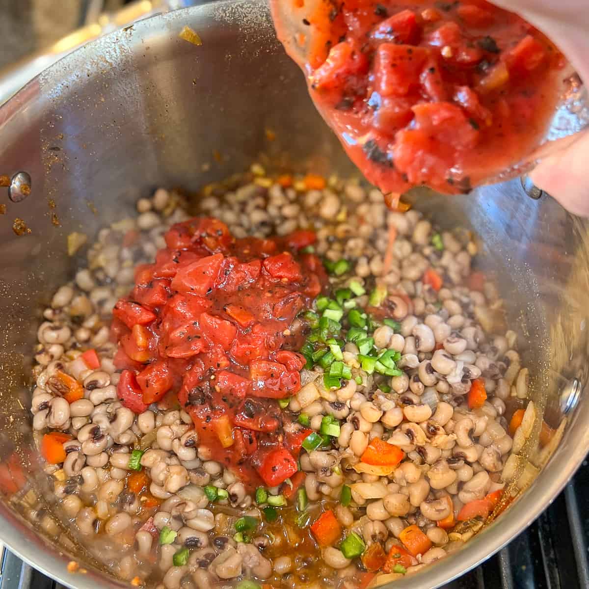 top side view of fire roasted diced tomatoes being add to a pot with cooked black eyed peas, chopped jalapenos and veggies