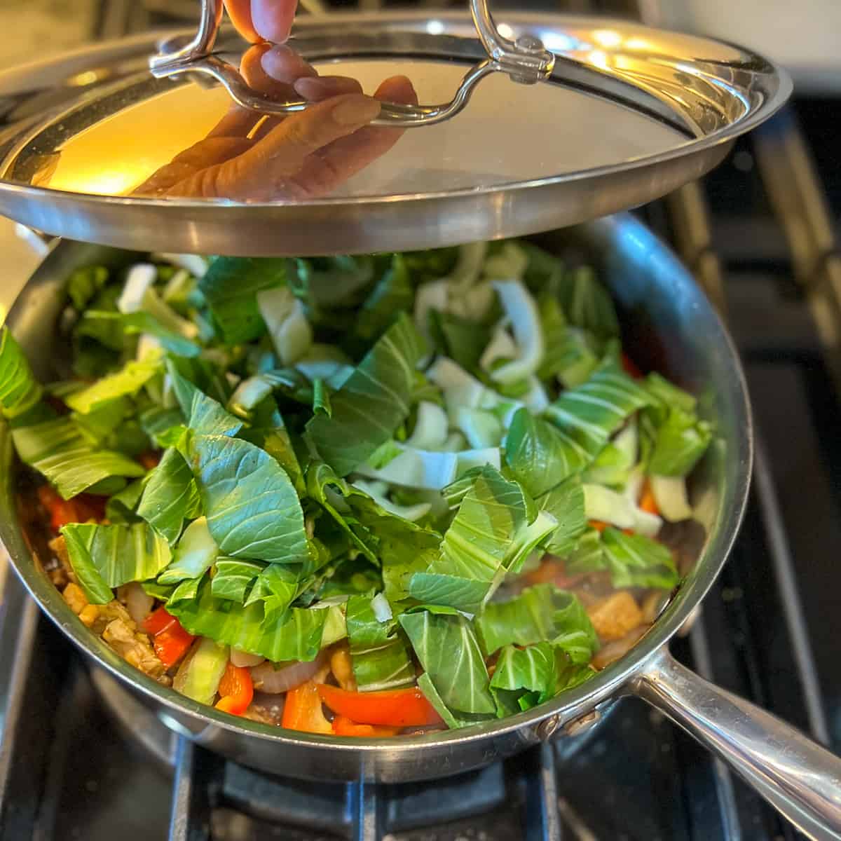 top side view of a lid being put onto a pan with chopped baby bok choy; underneath the baby bok choy is tempeh and other veggies