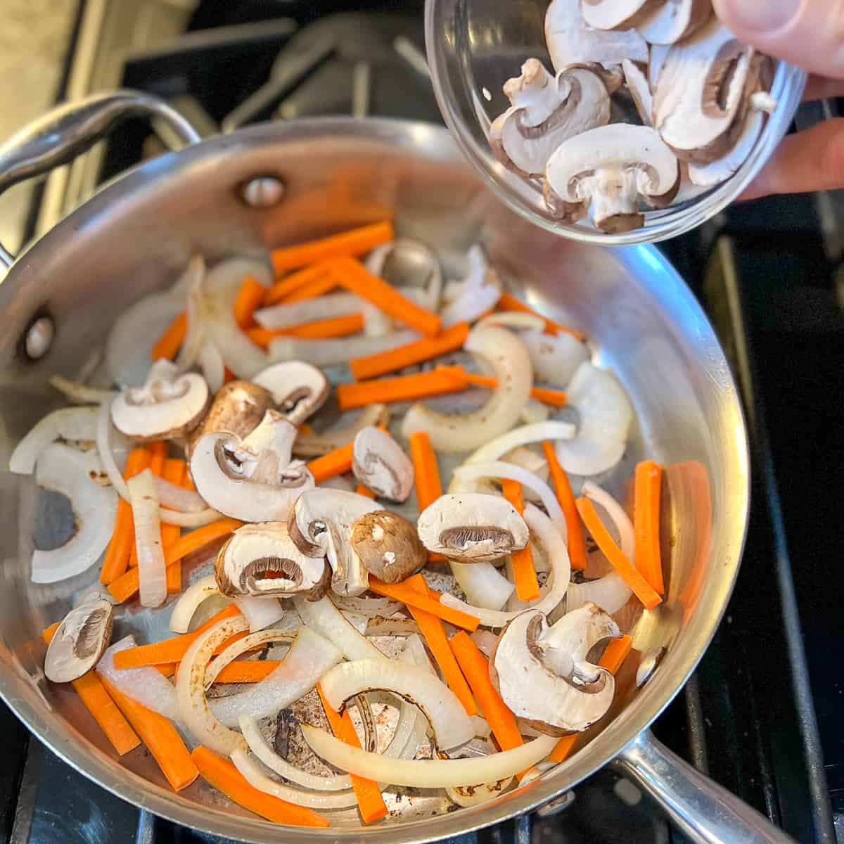 top view of sliced mushrooms being added to pan with carrots and onion