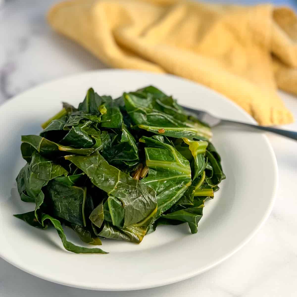 side top view of a plate of cooked vegan collard greens on white plate with fork and yellow napkin blurred in background