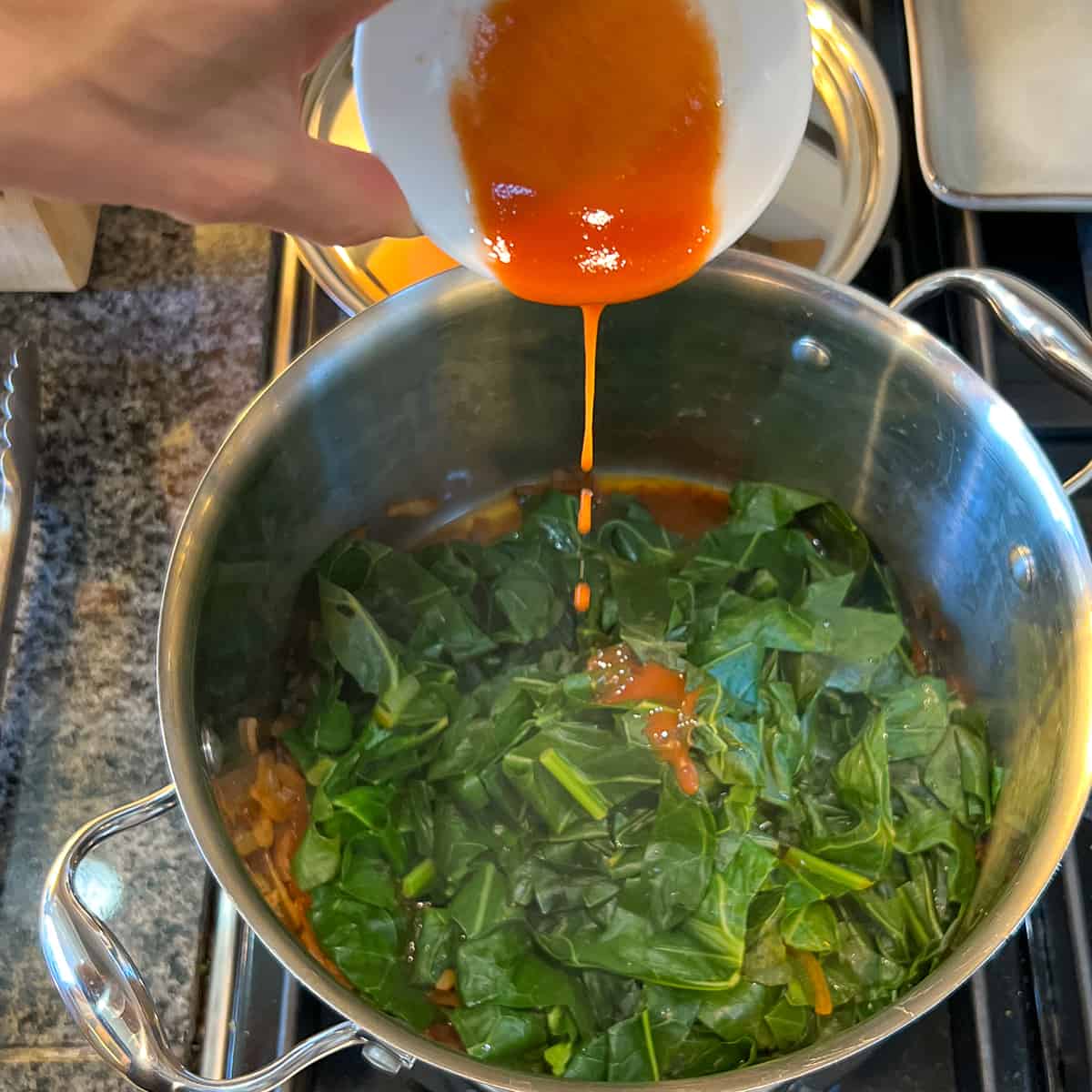 top view of large pot of cooked collard greens with red pepper hot sauce being added