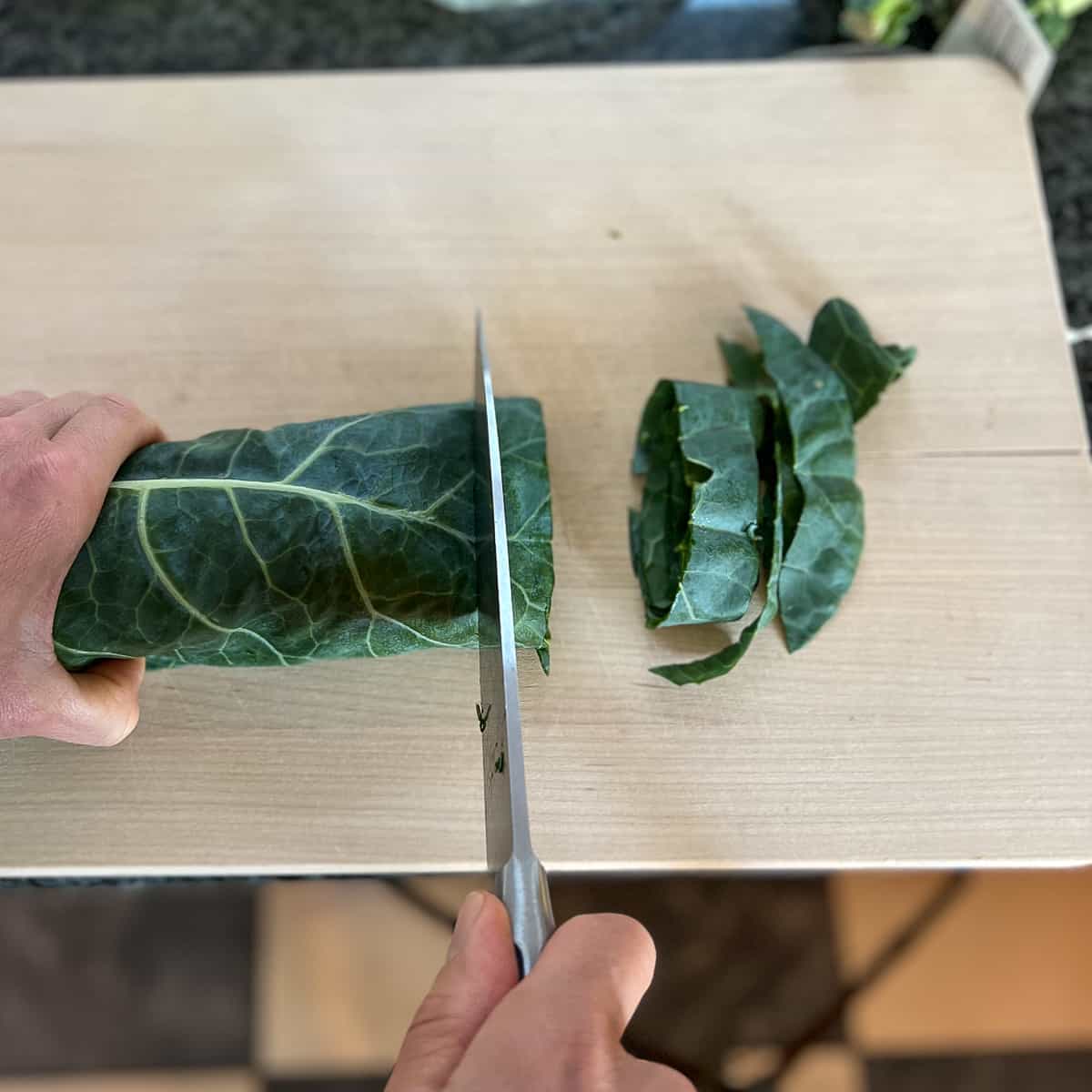 a woman's hand chopping collard greens into strips on a wooden cutting board
