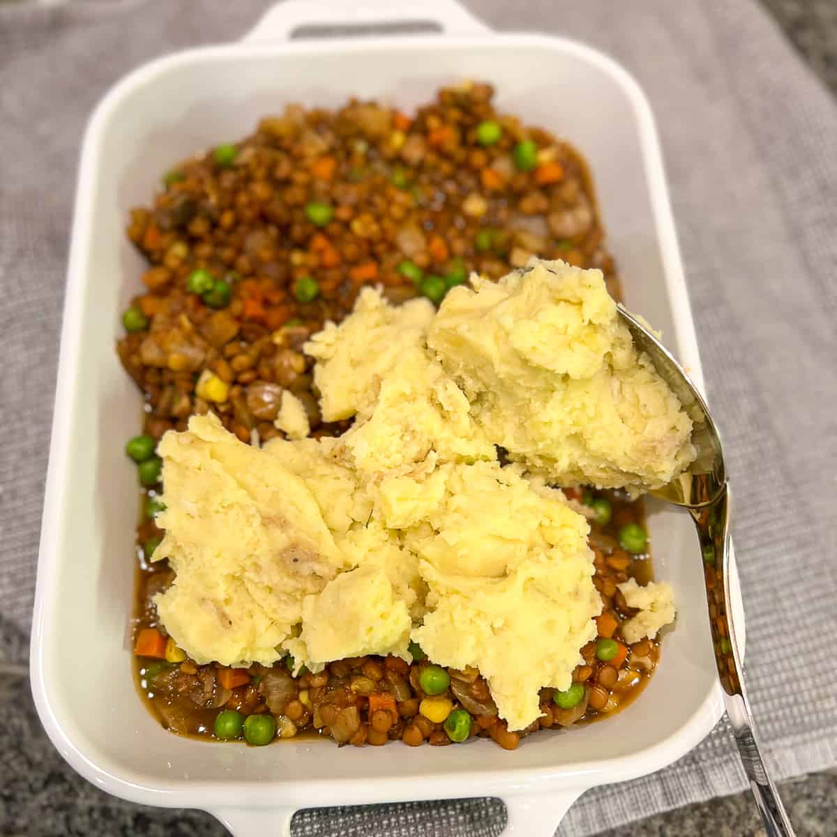 Lentil mixture in the bottom of the casserole dish with garlic mashed potatoes being added with a large spoon. 