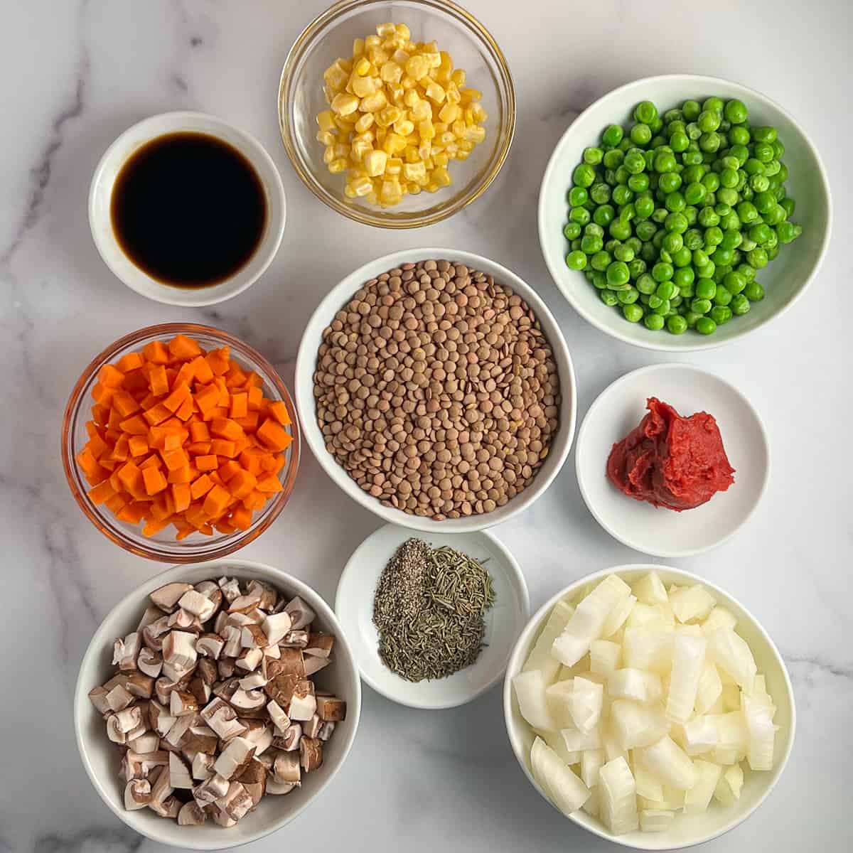 top view of key ingredients for vegan shepherd's pie: lentils, corn, peas, coconut aminos, diced carrots, tomato paste, diced mushrooms, chopped onions, seasonings