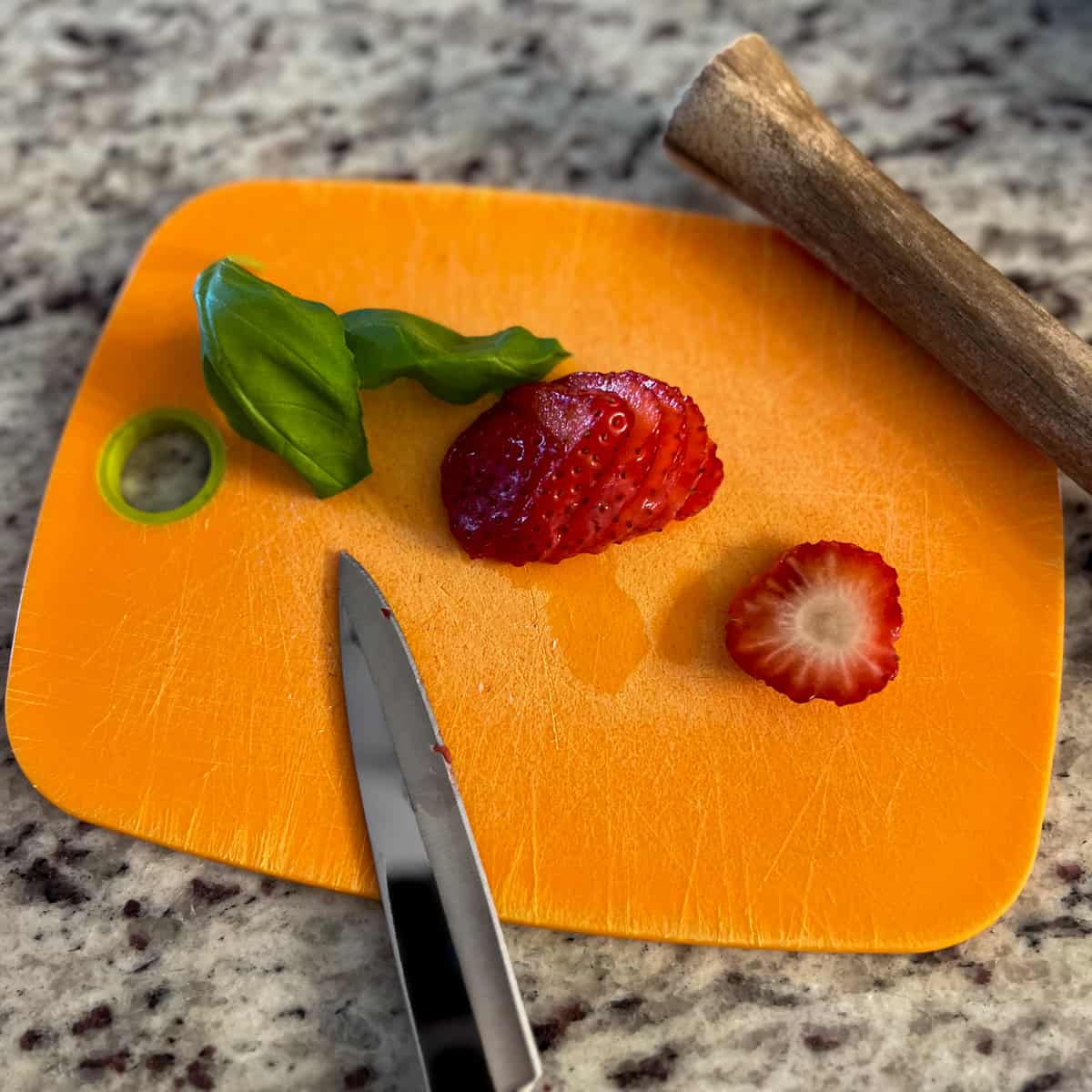 top side view of sliced strawberry, basil, knife and wooden muddle on a yellow cutting board