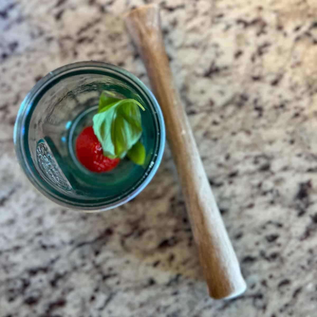 top view of a glass with fresh strawberry and basil leaves inside and wooden muddle on the side