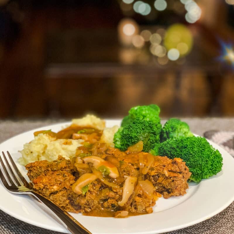 side view of lentil loaf, mashed potatoes with gravy and broccoli on a white plate; Christmas tree blurred in the background