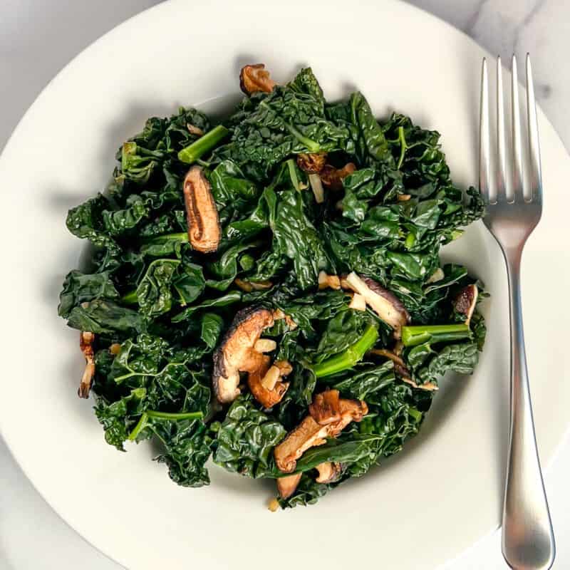 top view of sauteed kale and mushrooms with garlic in a white rimmed bowl with a fork on the side