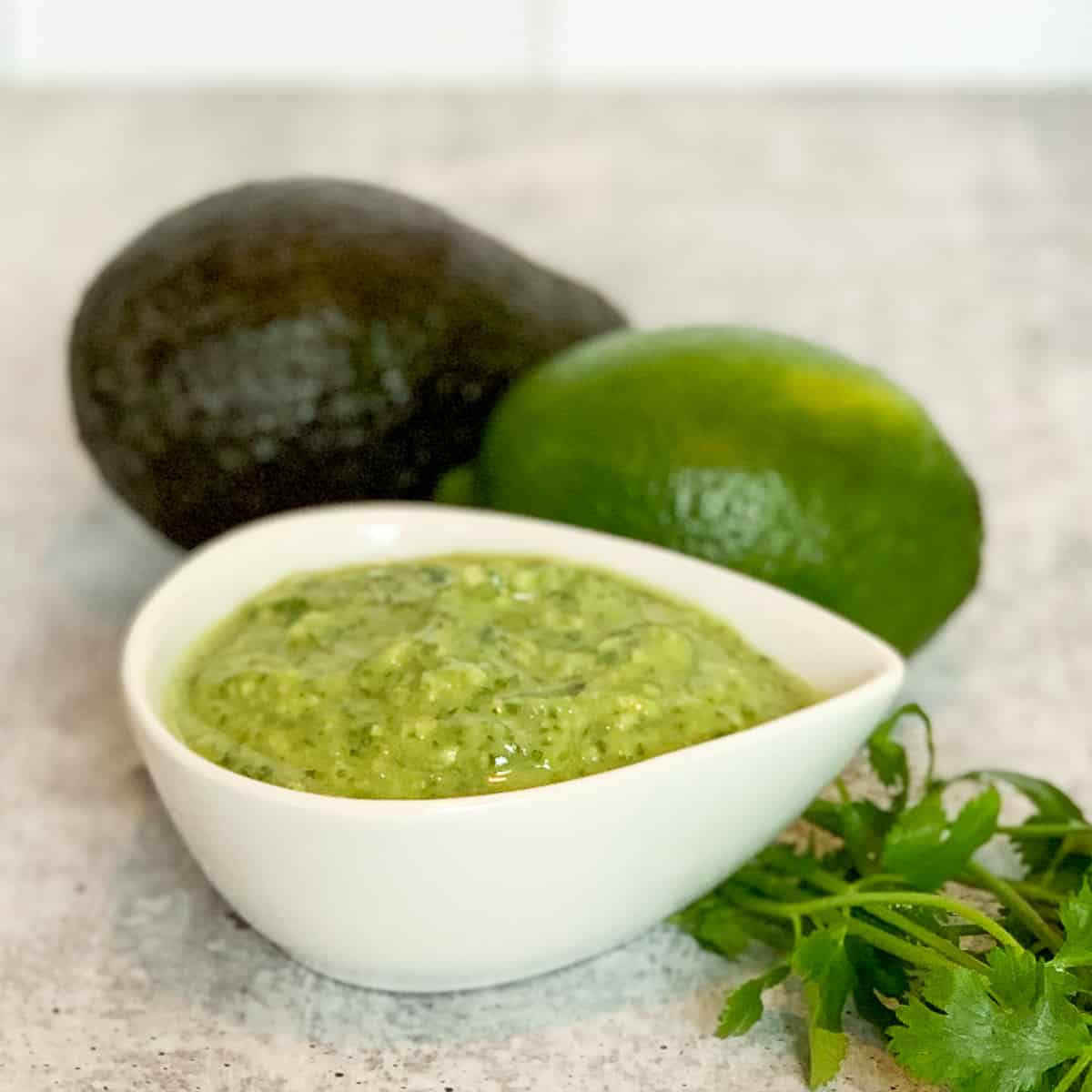 side view of avocado, cilantro lime dressing in a small white dish with fresh cilantro, lime, and avocado blurred in the background.