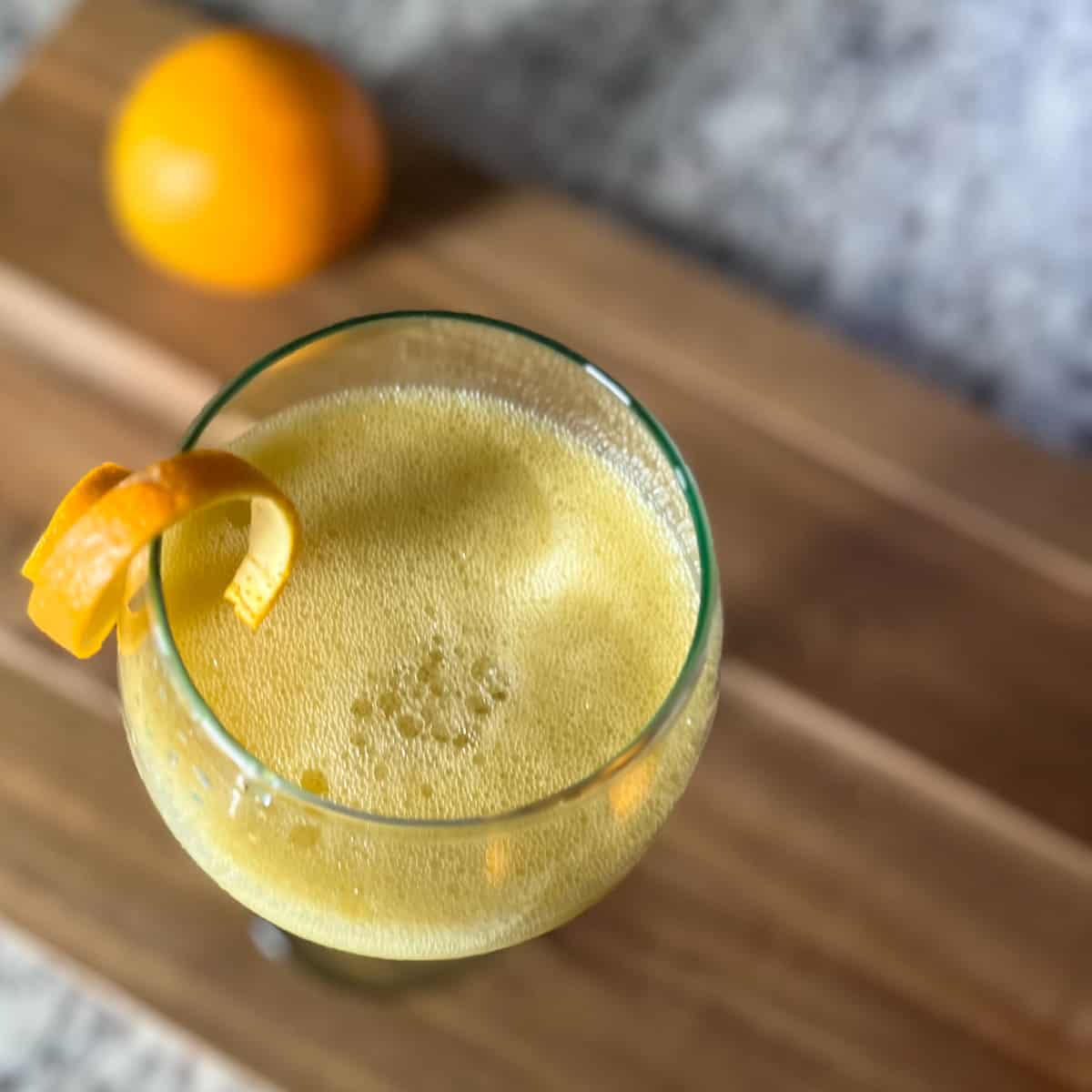 top view close up of orange juice mocktail in a stemmed glass on wooden cutting board; orange peel balancing on the rim of the glass; whole orange blurred in the background