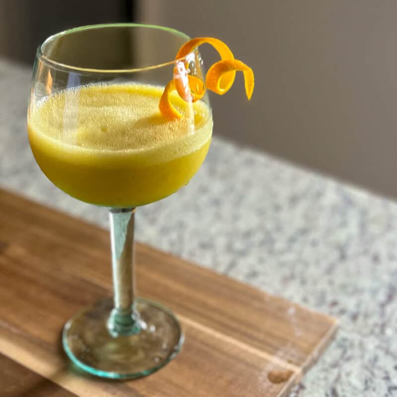 side view of orange juice mocktail in rimmed glass placed on a wooden cutting board; a twisted orange peel balancing on the rim of the glass