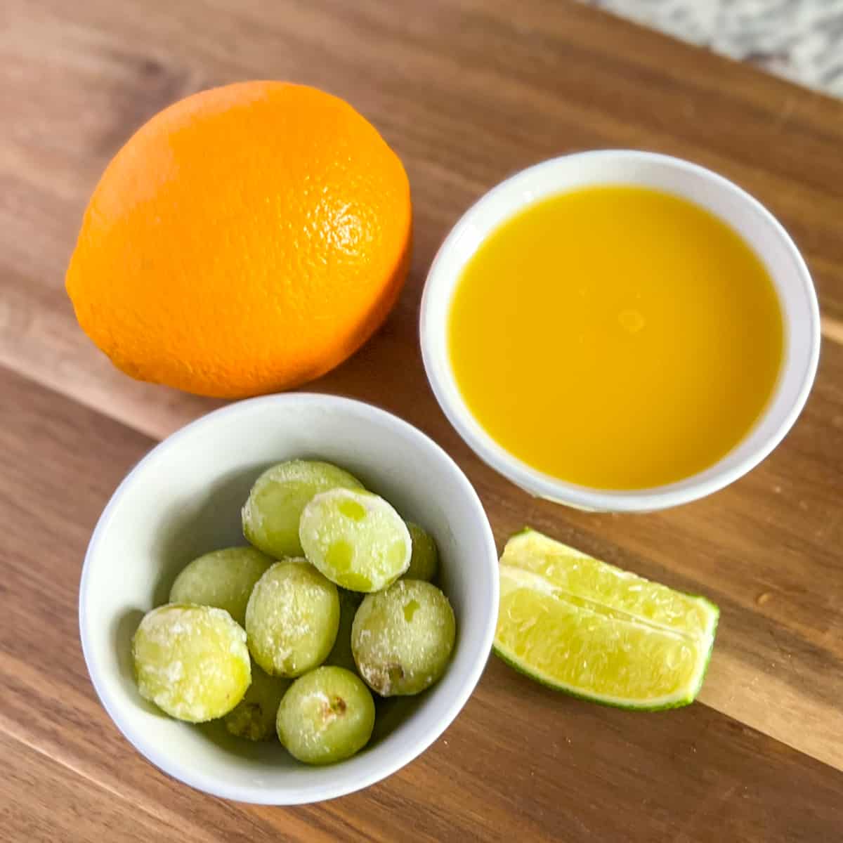 top view of ingredients on wooden cutting board used in orange juice mocktail: orange juice, fresh orange, frozen grapes, lime wedge