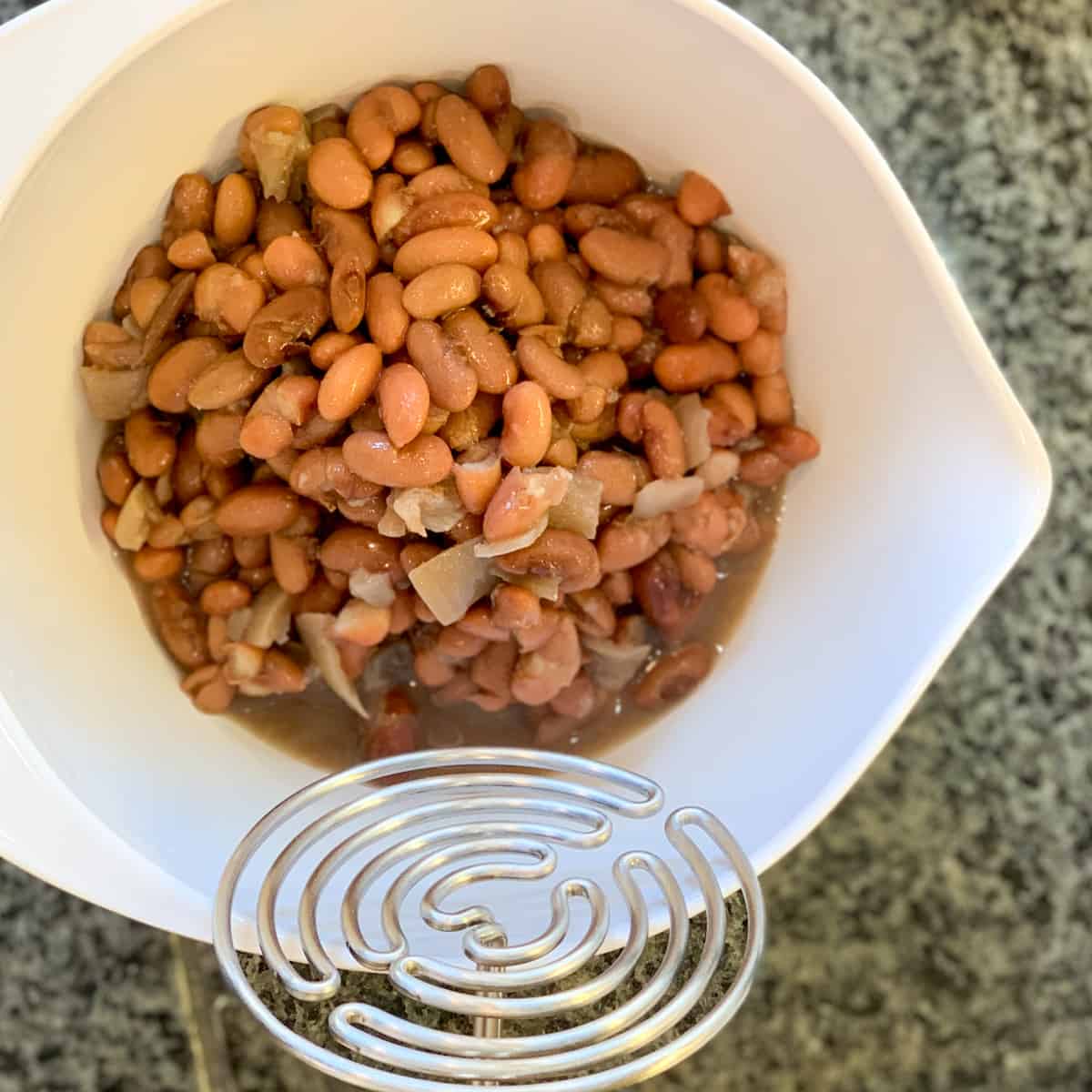 close up top view of cooked beans in a large white mixing bowl and potato masher to make the refried beans 