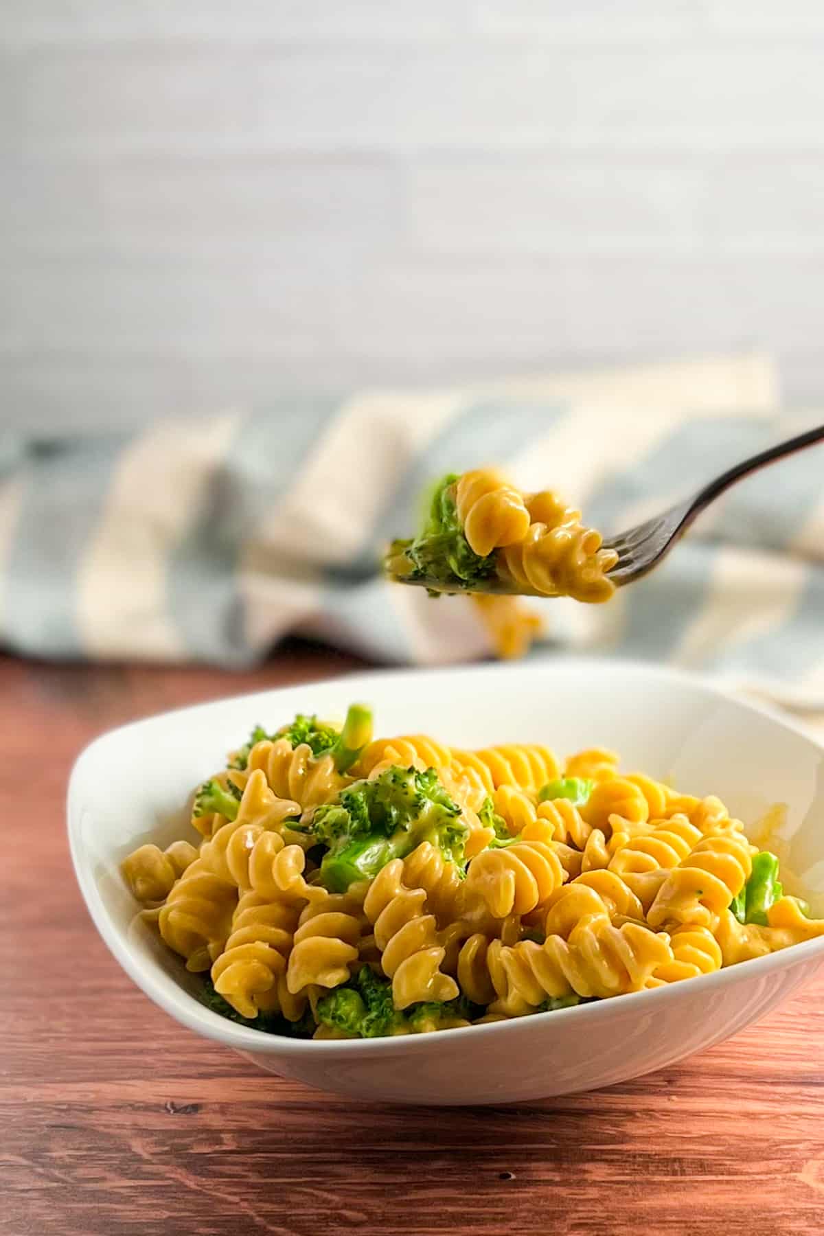 side view of vegan mac and cheese in a white bowl on a wood surface with a fork suspended above the bowl. blue and white striped napkin blurred in the background