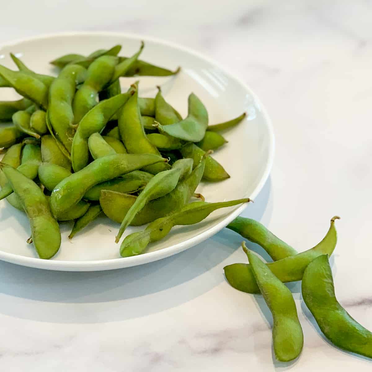 side close up of cooked edamame in the pods on a white plate and a few spilling over onto a marble surface