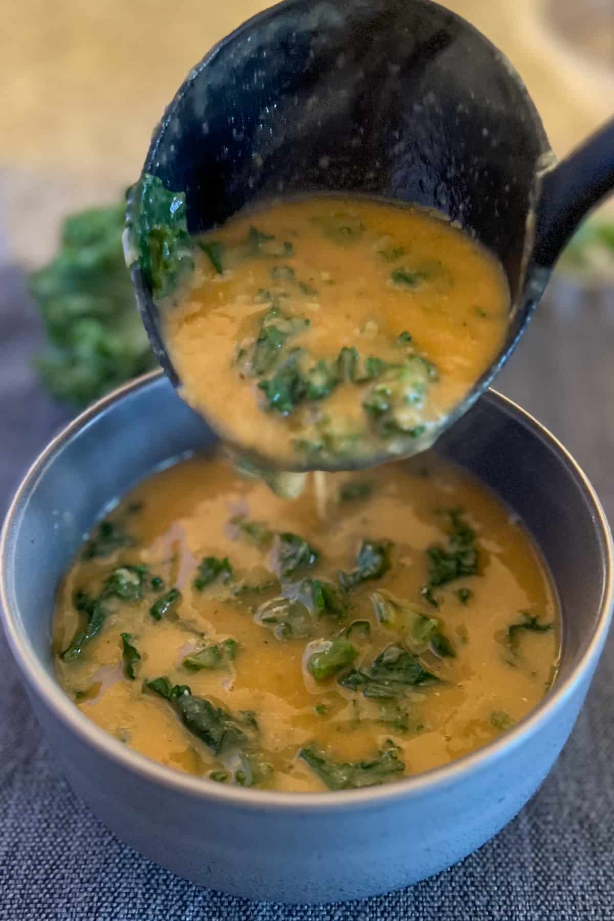 top side view of a blue bowl with white bean and kale soup; black ladle is pouring more hot soup into the bowl