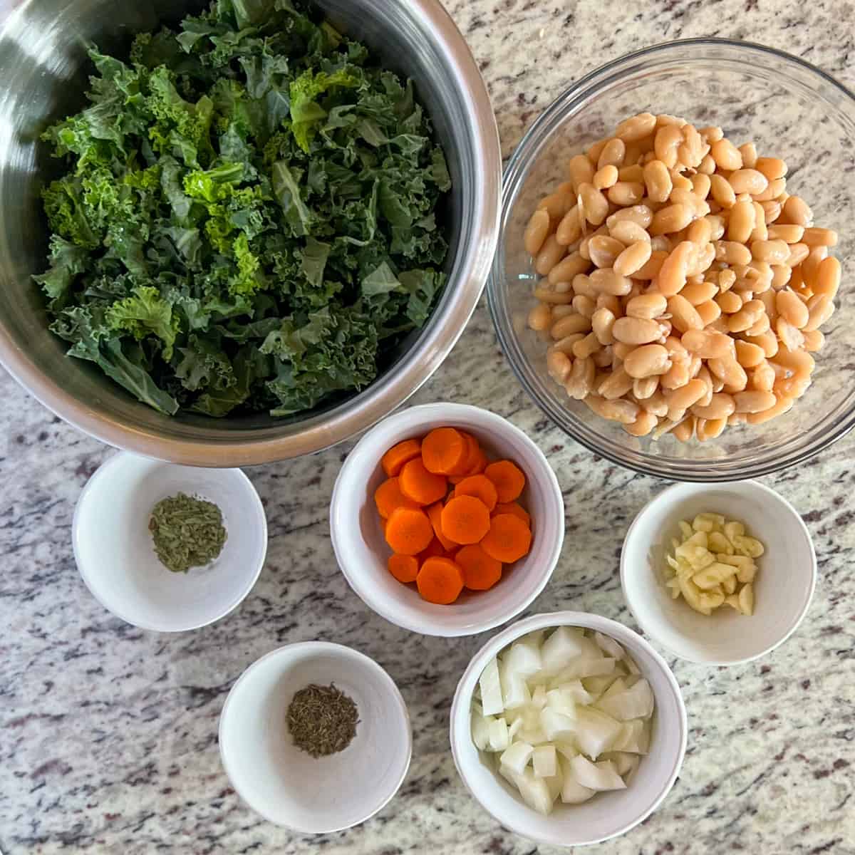 top view of key ingredients used in the recipe: cooked cannellini beans, chopped kale, onion, carrot, garlic, thyme, and tarragon