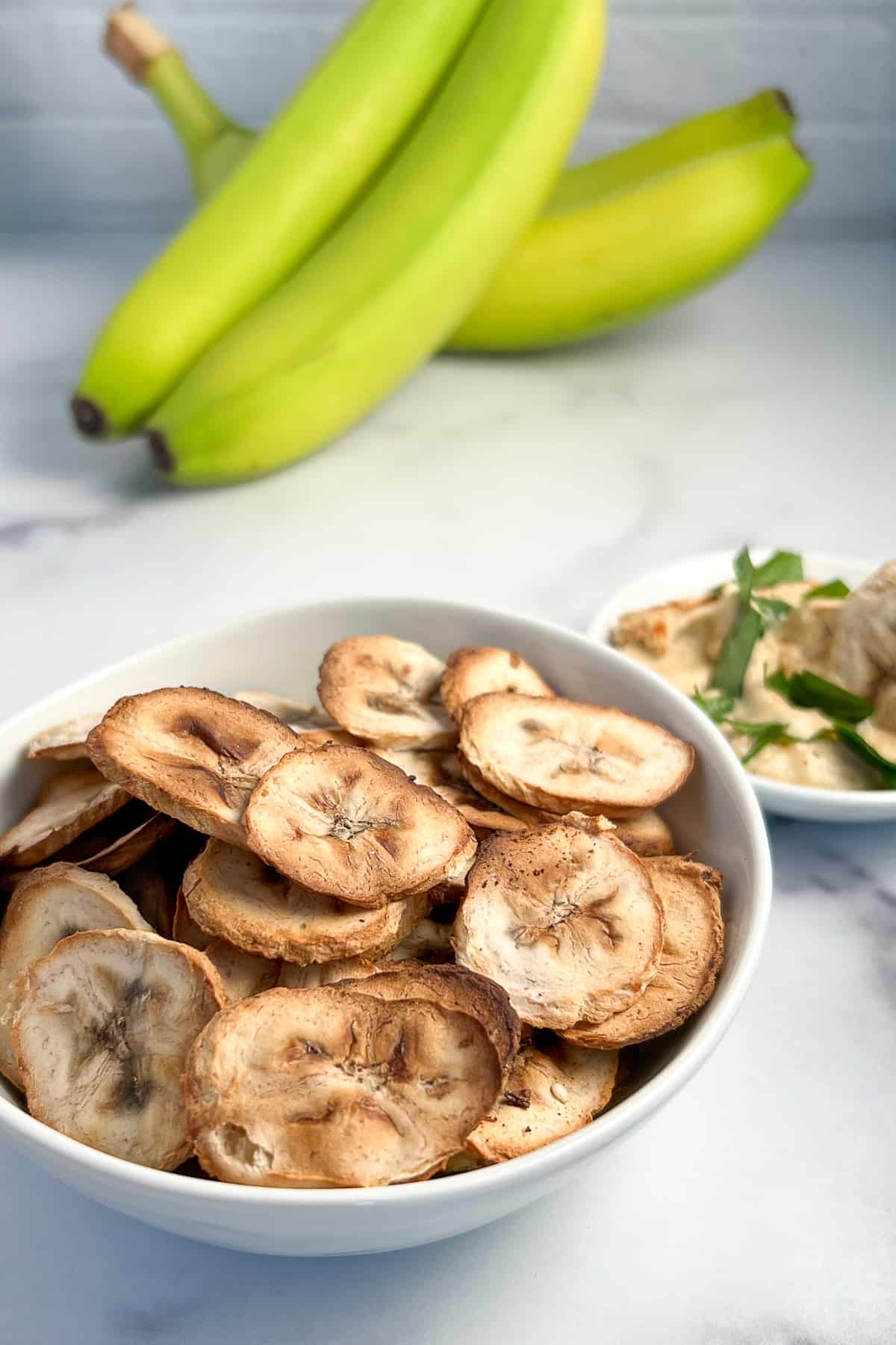 bowl of oil free savory air fryer banana chips in a white bowl with green bananas and a small bowl of hummus with fresh parsley blurred in the background