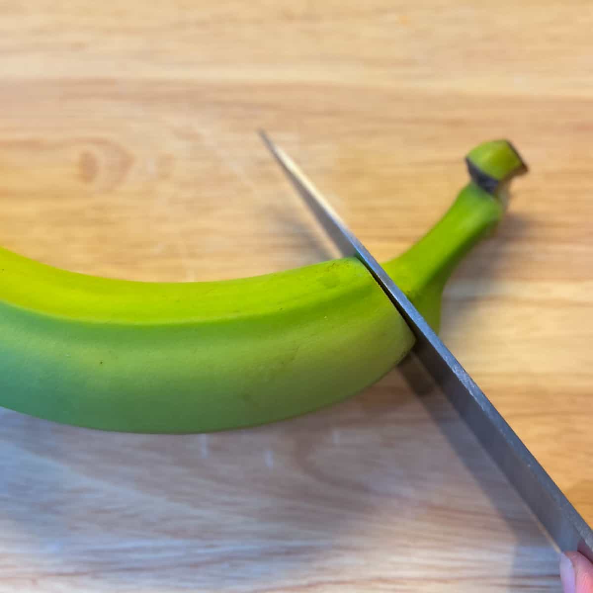 top view of a knife cutting the stem off of a green banana on a cutting board