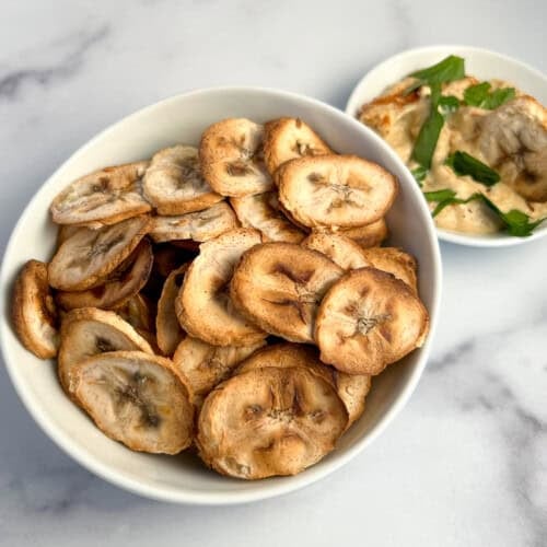 top side view of a bowl of banana chips with hummus and parsley blurred in the background