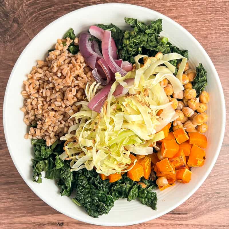 top view of baked salad with kale, cabbage, chickpeas, winter squash, farro and onions in a white bowl