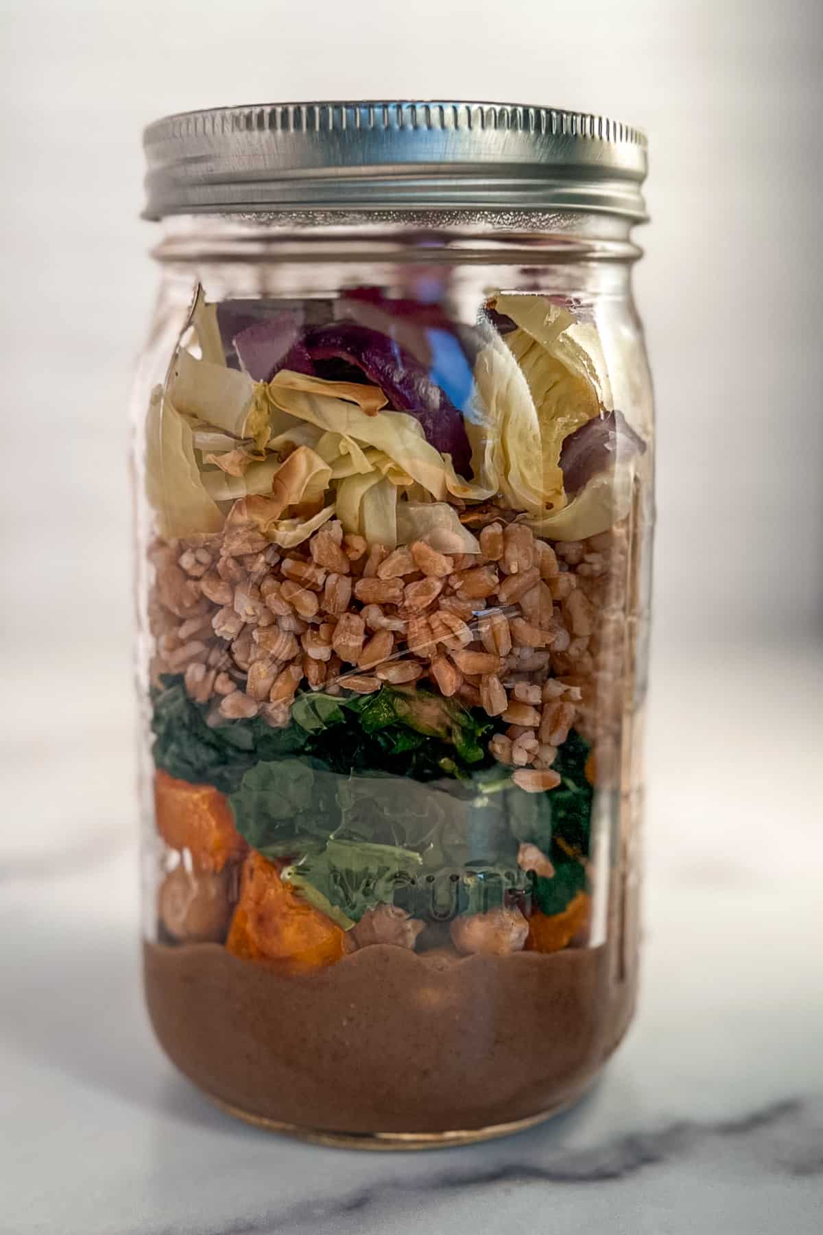 side view close up of baked salad in a mason jar with dressing on the bottom and layers of veggies, farro and chickpeas