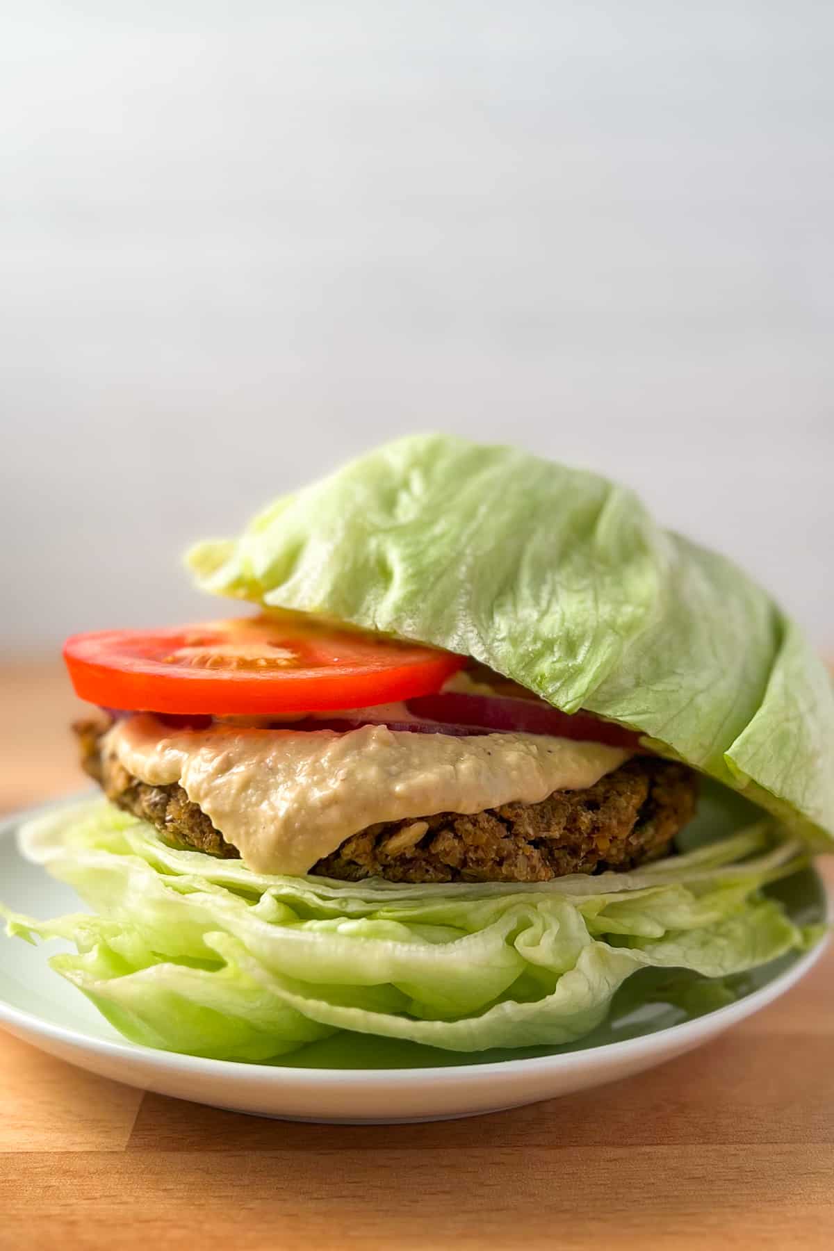 side view close up of chickpea burger with lettuce bun topped with sliced tomato, onion and hummus sauce