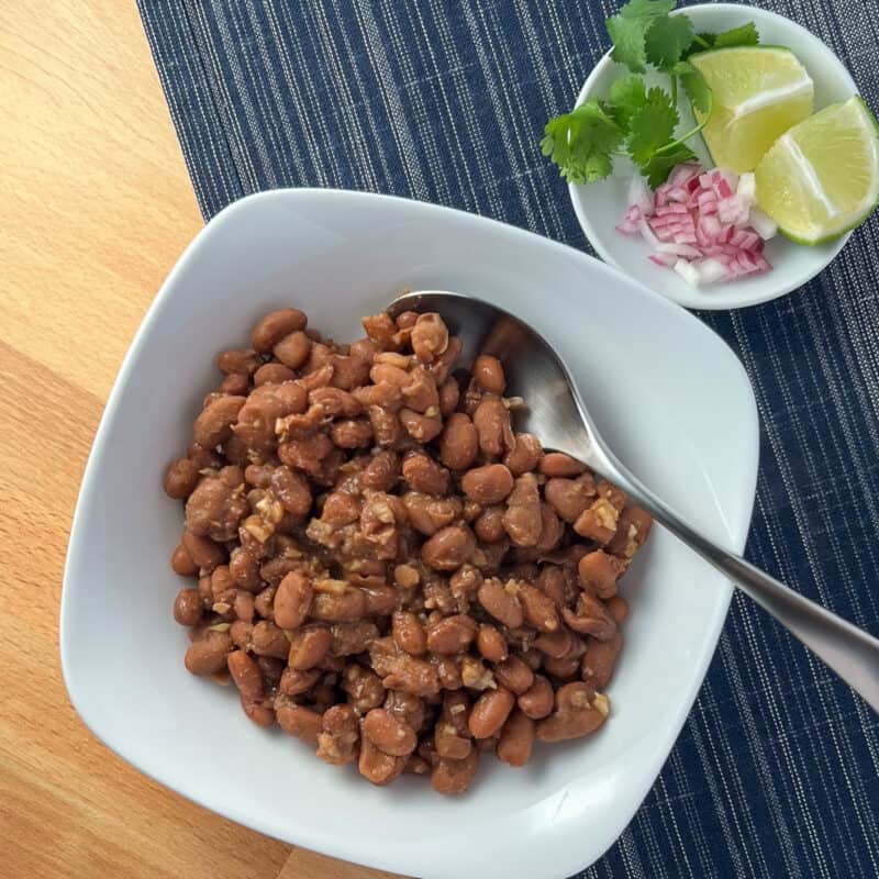 top view of chipotle pinto beans in a white bowl with diced red onion, fresh cilantro and lime on the side