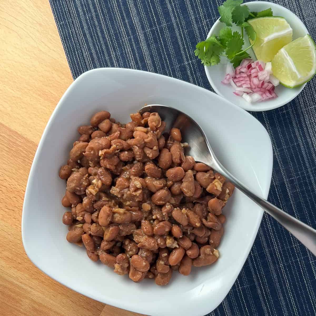 top view of a bowl of chipotle pinto beans with spoon; small dish with diced onion, fresh cilantro and lime wedges on the side