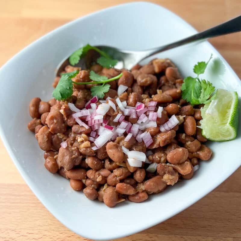 top side view of a bowl of chipotle pinto beans with spoon; small dish with diced onion, fresh cilantro and lime wedges on top