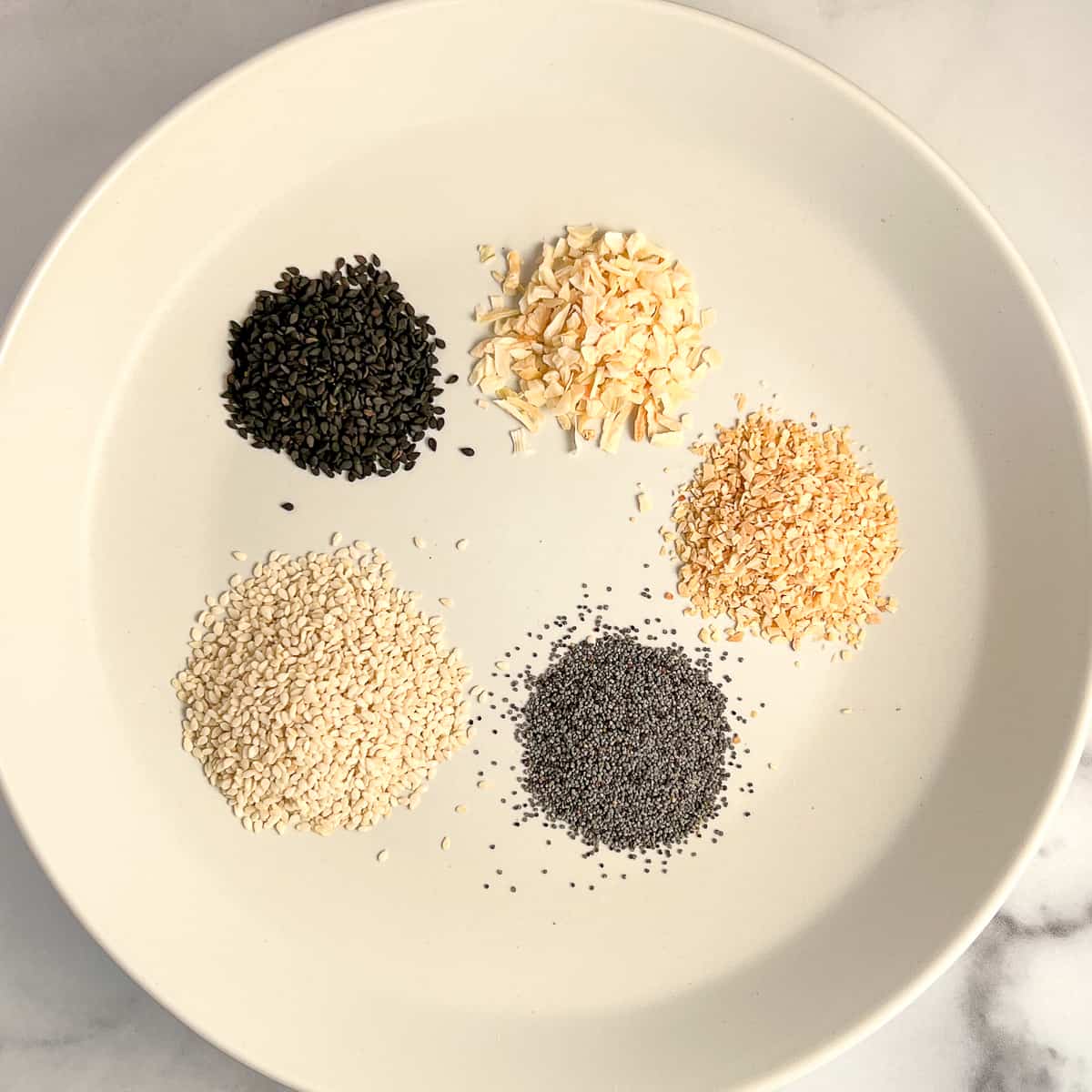 top view of a white plate with separated seasonings: black sesame seeds, white sesame seeds, poppy seeds, minced dried garlic, minced dried onion