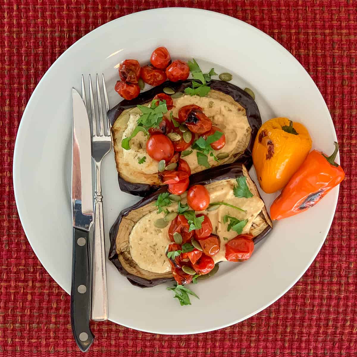 top view of two eggplant steaks topped with hummus, grilled tomatoes, pumpkin seeds and fresh chopped parsley; grilled mini bell peppers and steak knife and fork on the side