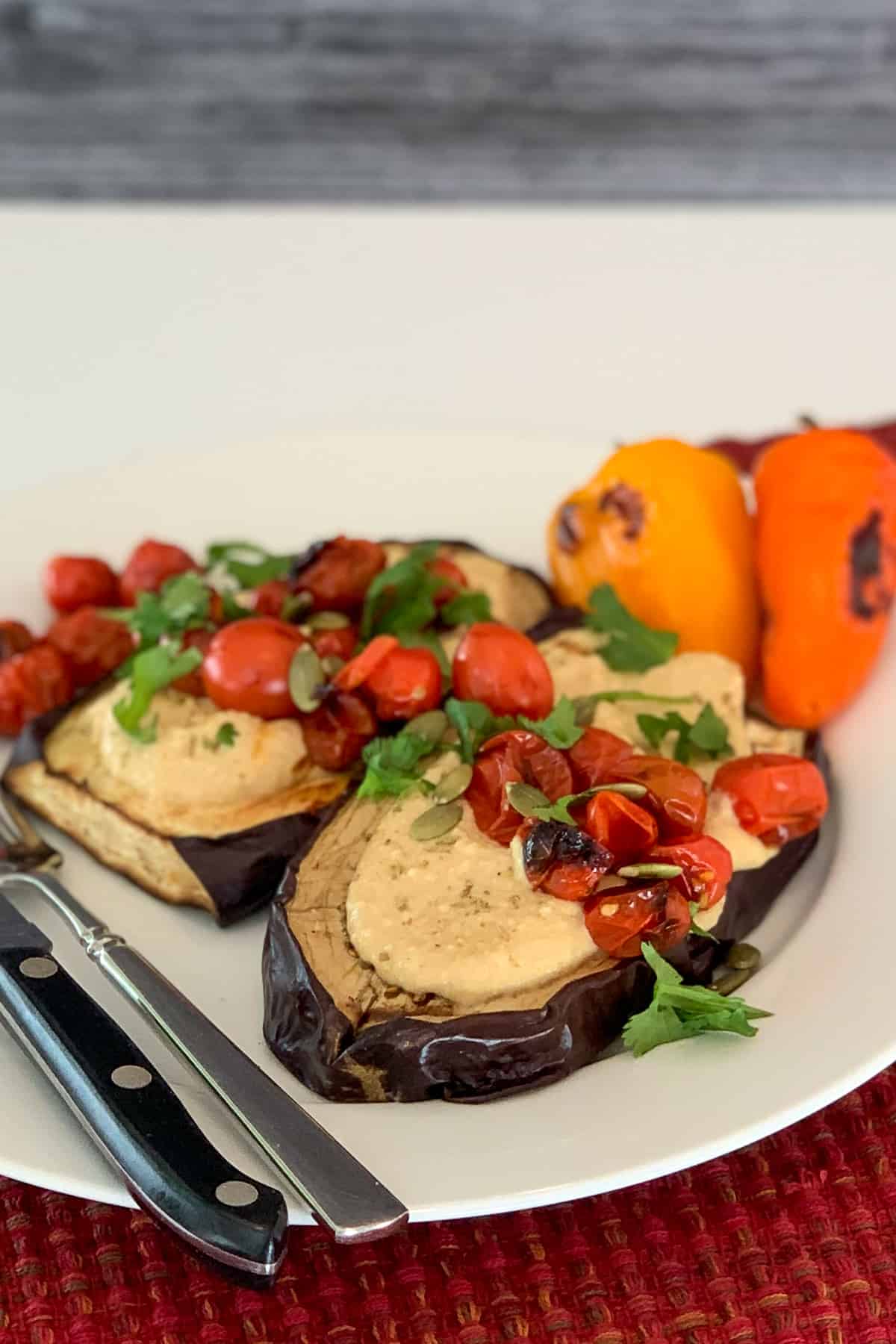 top side view of two grilled eggplant steaks topped with hummus and grilled cherry tomatoes, pumpkin seeds and fresh parsley; grilled mini bell peppers blurred on the back side of the plate