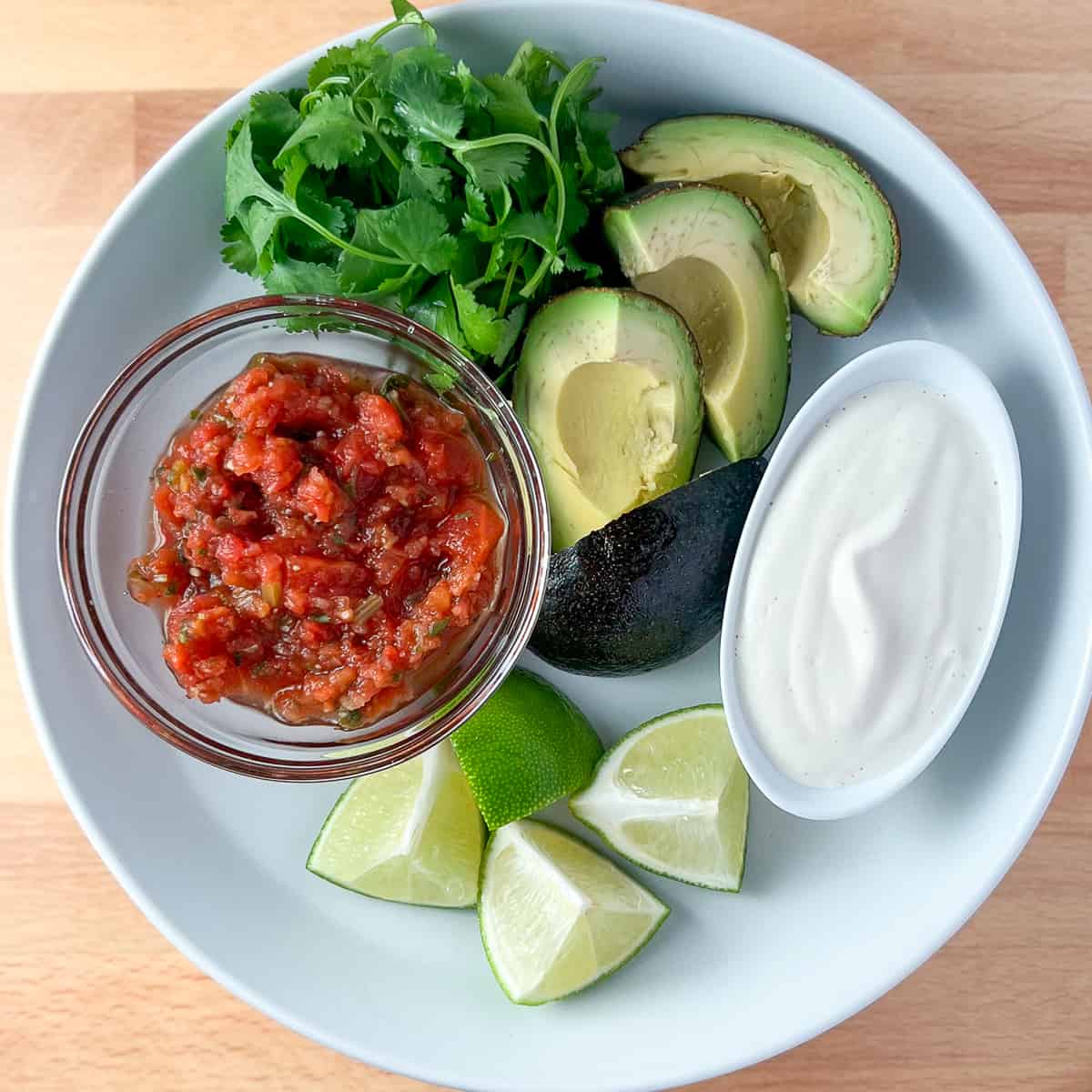 top view of what to add to vegan fajitas: salsa, fresh cilantro, sliced avocado, cashew cream and fresh lime wedges